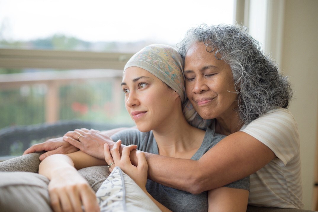 Mujer con pañuelo rodea afectuosamente a su madre en el sofá, mostrando apoyo en su lucha contra el cáncer.