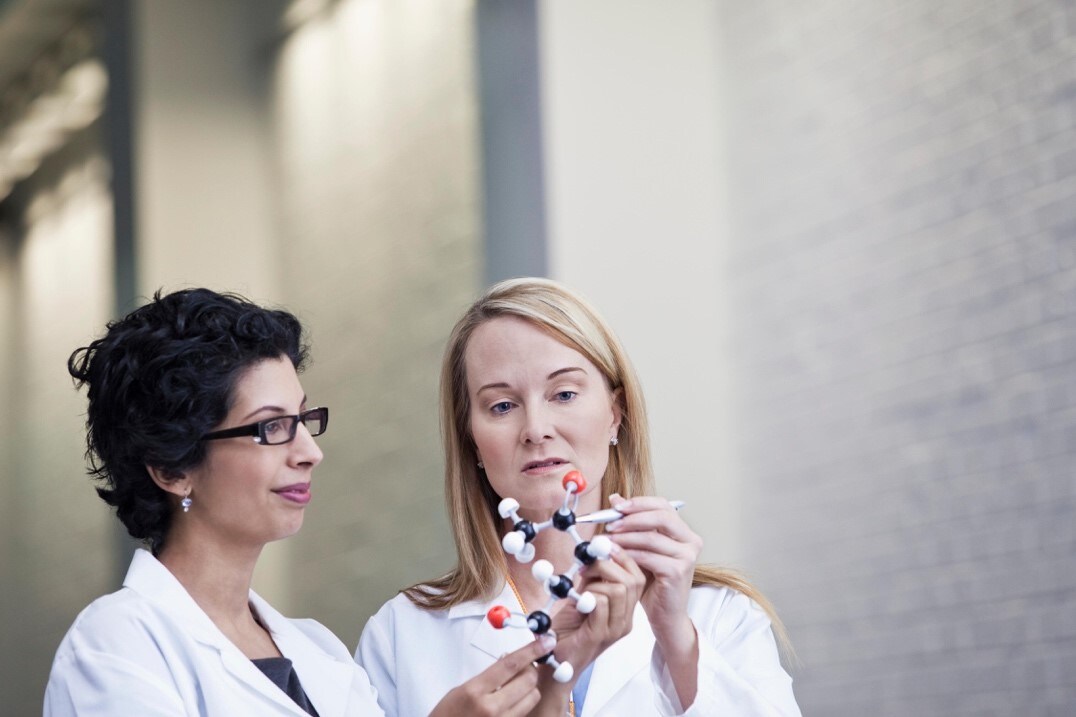 Dos mujeres en batas de laboratorio presentan un modelo molecular, destacando su trabajo.